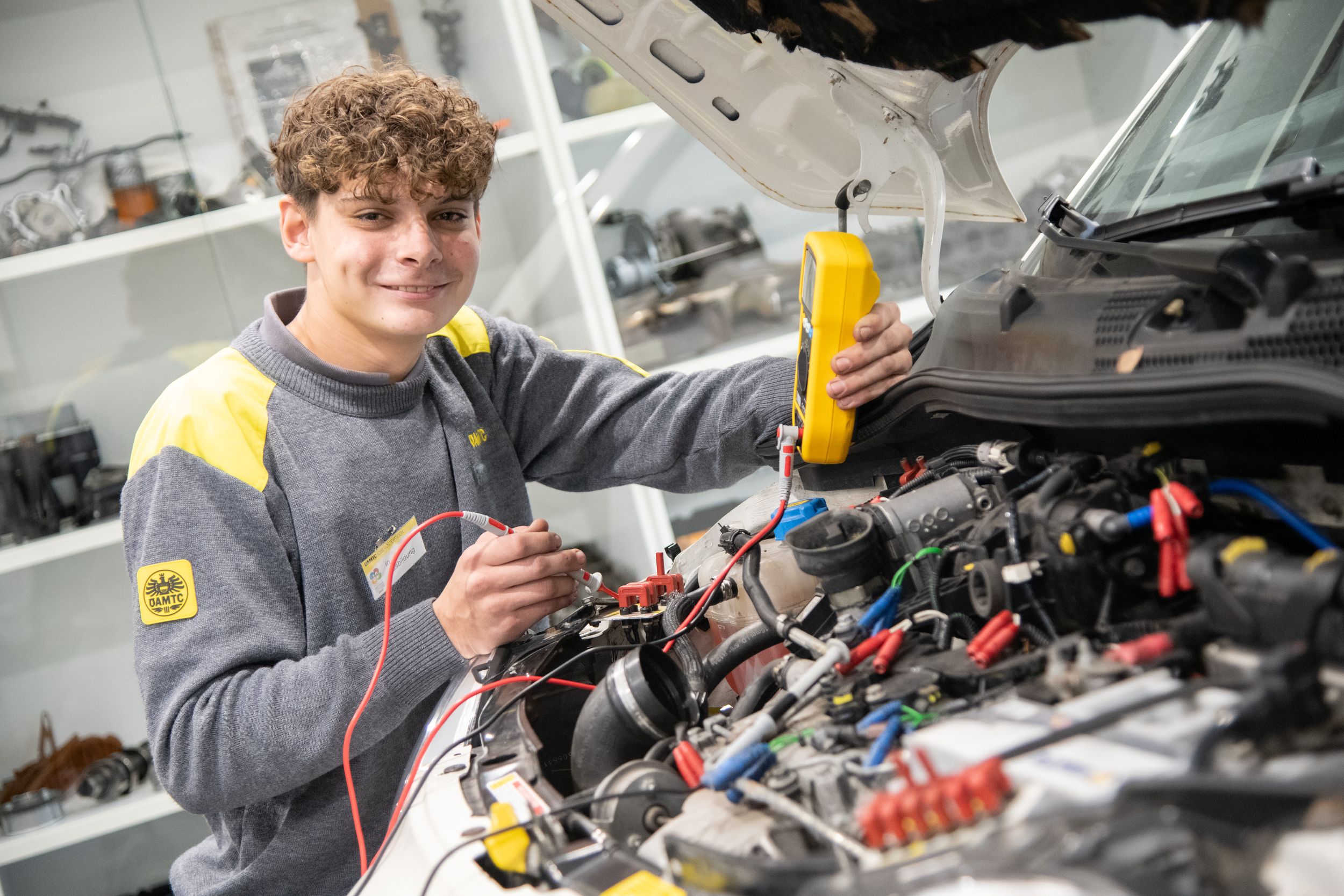 Lehrling mit Messinstrument in der Hand, neben offener Motorhaube